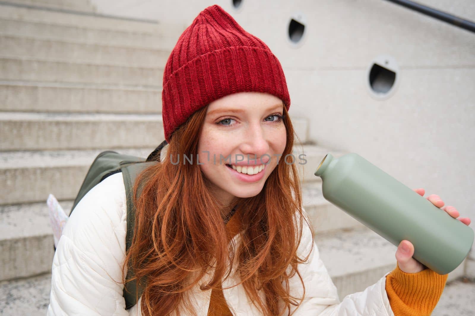 Happy redhead woman, tourist with thermos, drinks her hot tea, coffee from travel flask, restests during her travelling in city and sightseeing.
