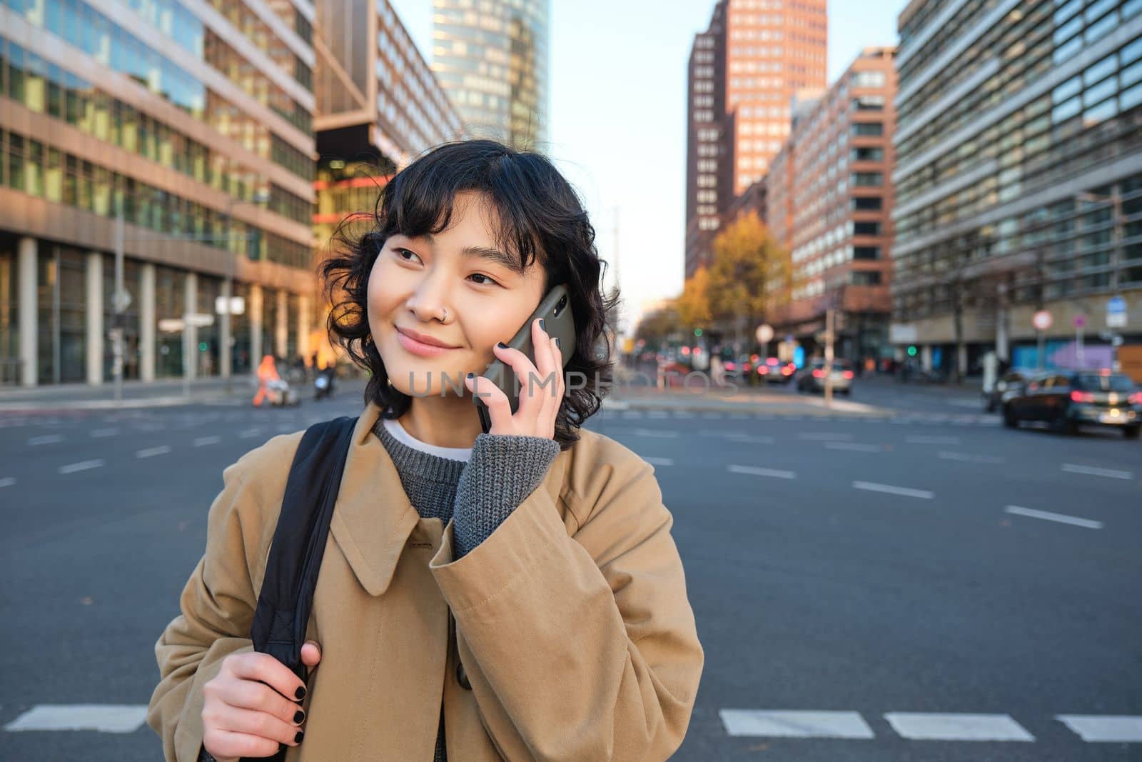 Smiling asian girl makes a phone call, stands on an empty street, calling someone on telephone, waiting for friend in city, going to a meeting by Benzoix