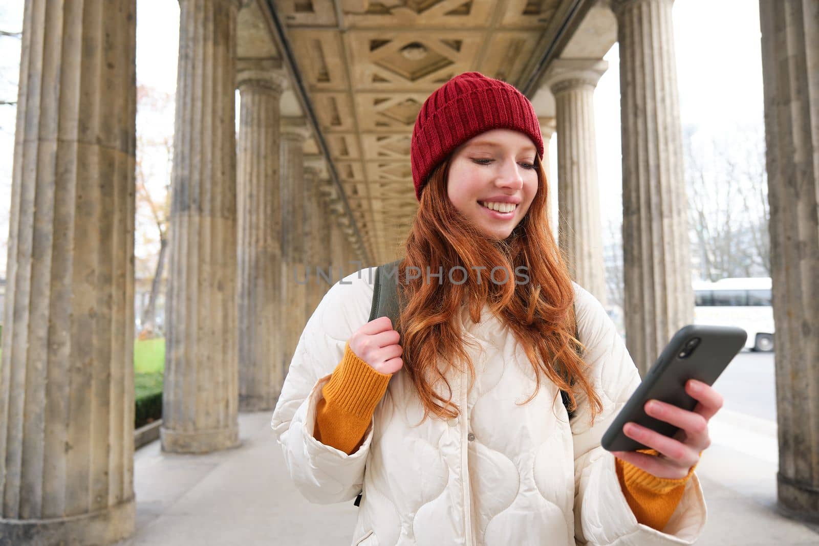 Mobile broadband and people. Smiling redhead 20s girl with backpack, uses smartphone on street, holds mobile phone and looks at application.