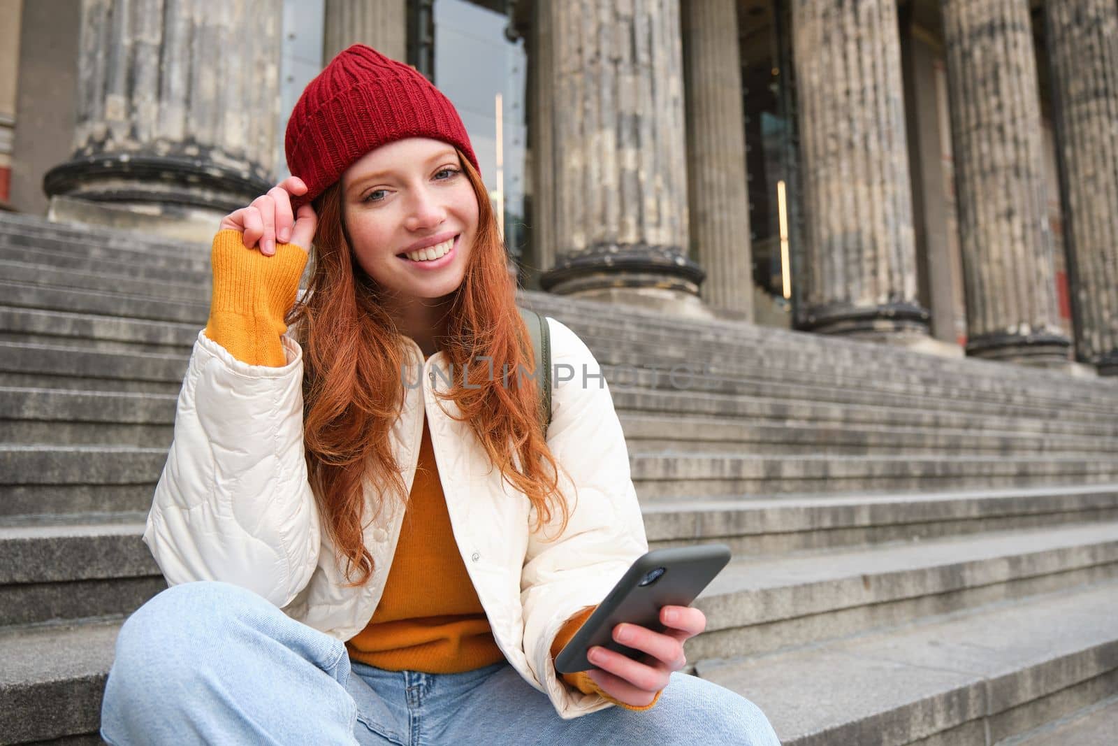 Portrait of young urban girl in red hat, sits on stairs near museum, holds mobile phone, connects to public wifi and surfs net, uses smartphone apps.