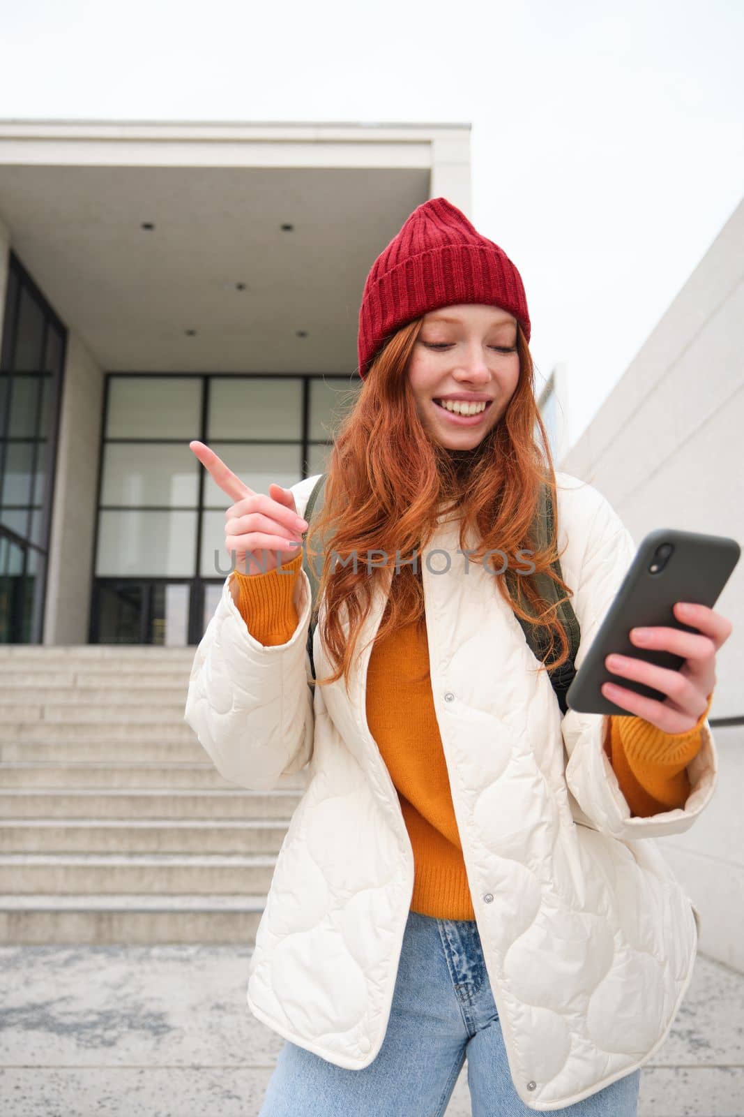 Vertical shot of redhead girl tourist, shows her friend via video chat building, points somewhere, talks on mobile phone.