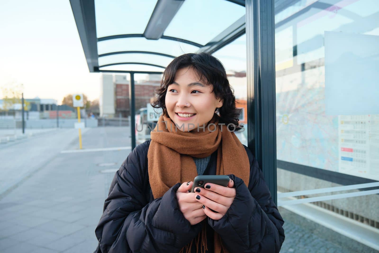 Portrait of Korean woman in winter jacket, standing with smartphone, waiting for bus on stop, looking at mobile phone app checking public transport application.