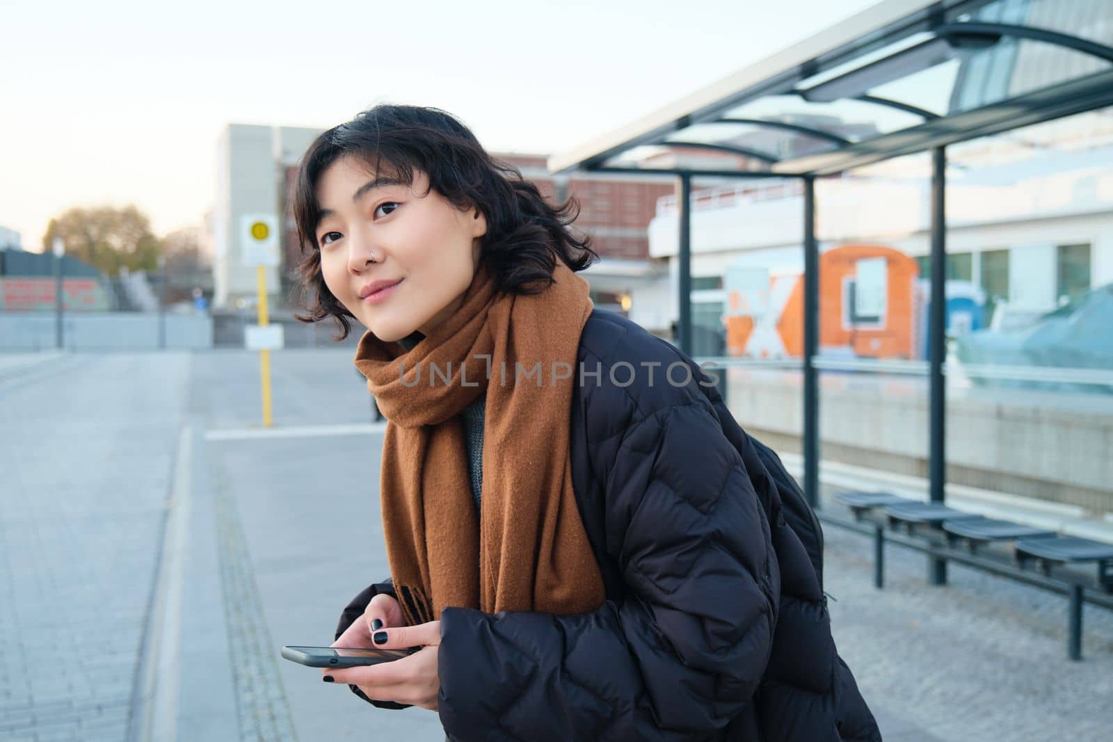 Cute smiling woman standing on a bus stop and looking at road, waiting for her public transport, holding smartphone, checking schedule on mobile app.