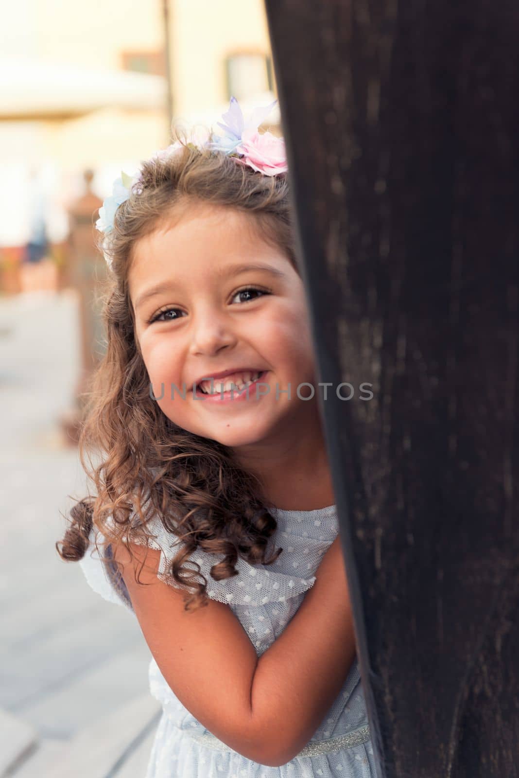 little girl smiles playfully while hiding, wearing a blue dress and a flower headband