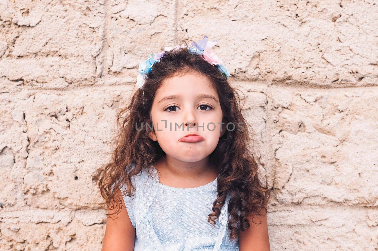 little girl making teasing with sad face, is in front of a stone wall and wears a blue dress and a flower headband