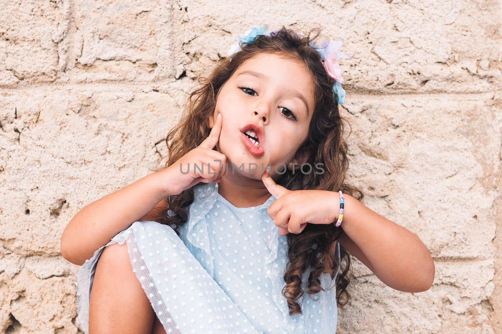 little girl making fun with hands and face, is in front of a stone wall and wears a blue dress and a flower headband