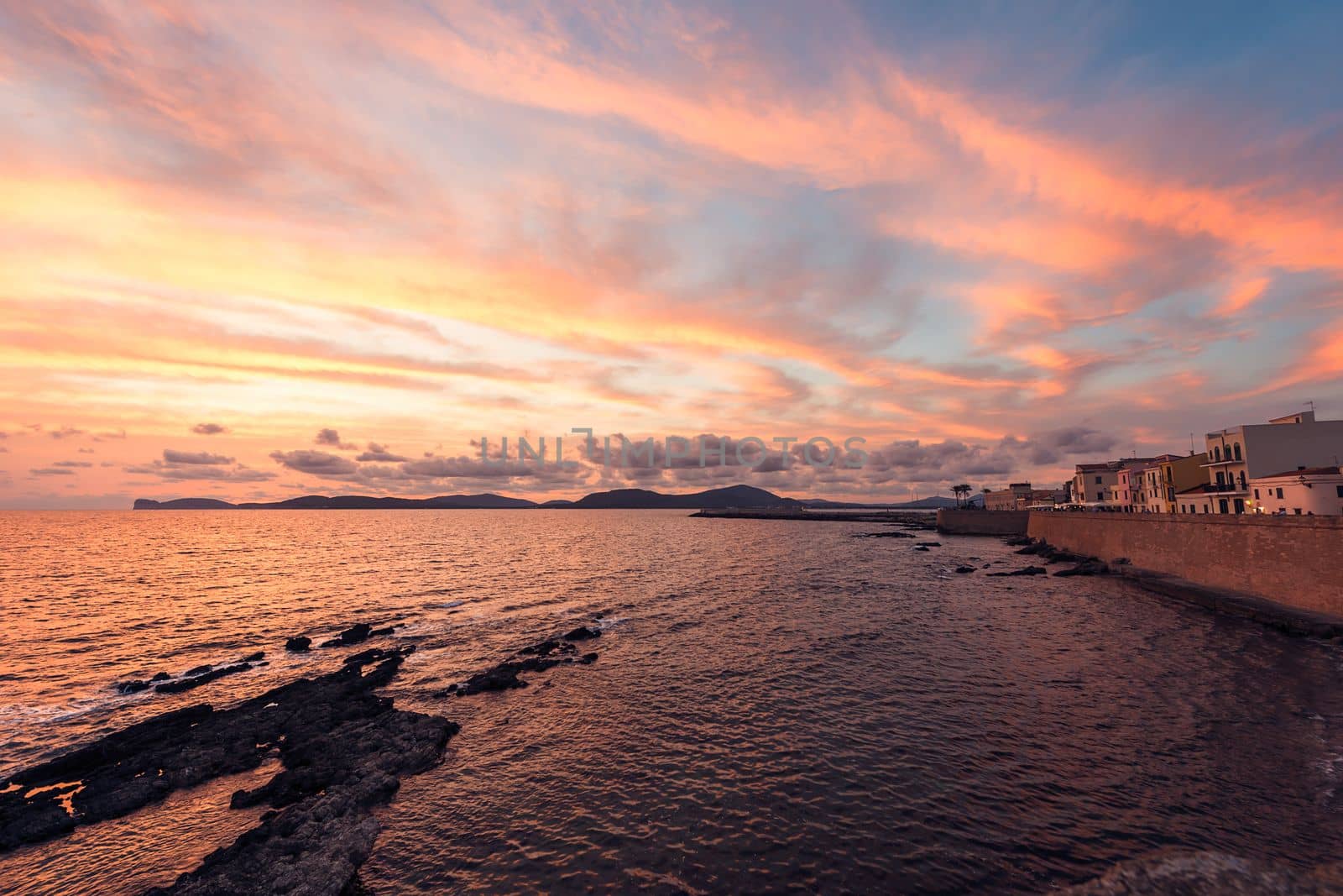 Spectacular and colorful sunset in Alghero, Sardinia, Italy. On one side is the seafront of the city and in the background the cape of the giant that sleeps