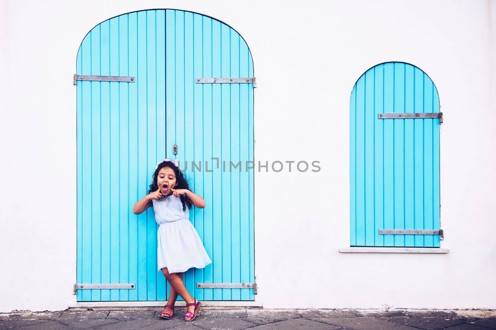 funny little girl making fun with her mouth, she is in front of a white wall with a door and a turquoise wooden window