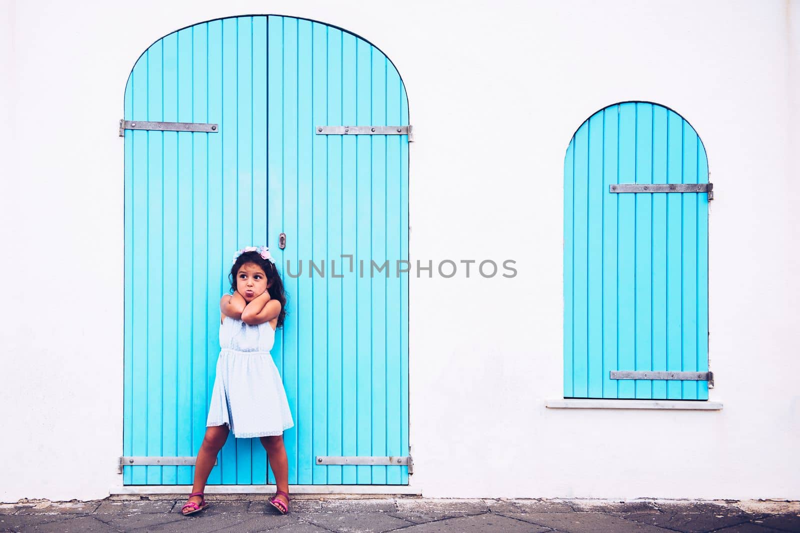 funny little girl making fun by sticking out her tongue, she is in front of a white wall with a door and a turquoise wooden window