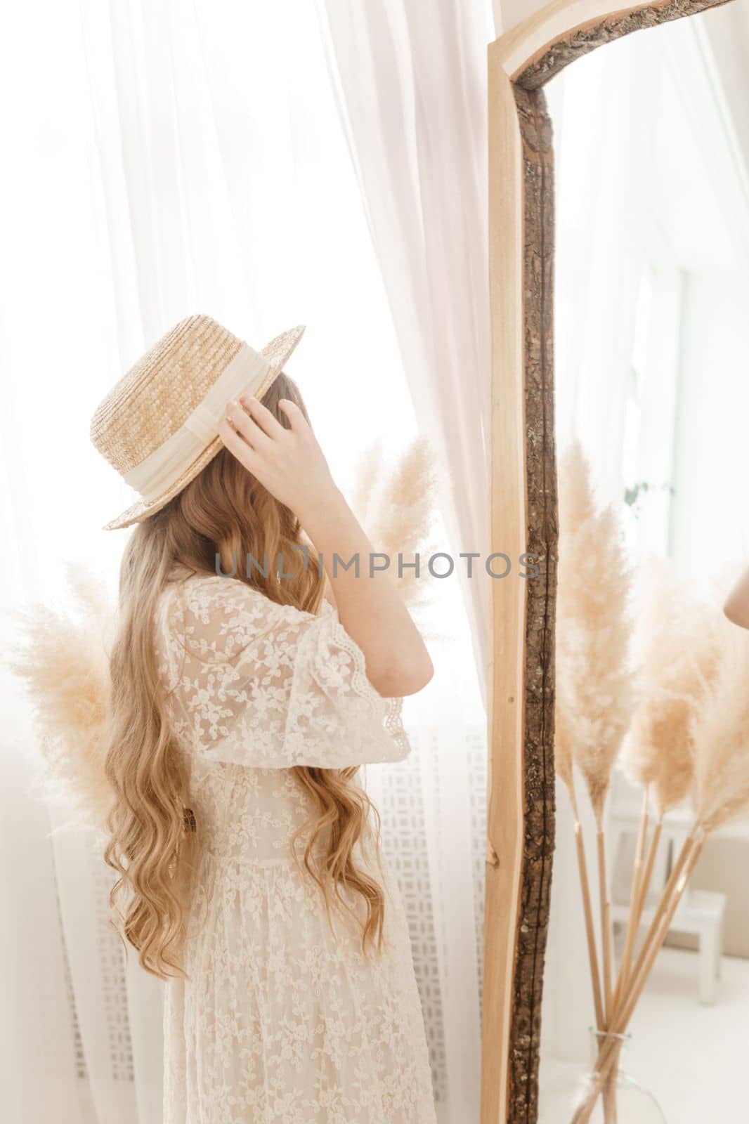 A beautiful teenage girl with long hair measures a straw hat in front of a mirror. Self-admiration of a blonde. selective focus
