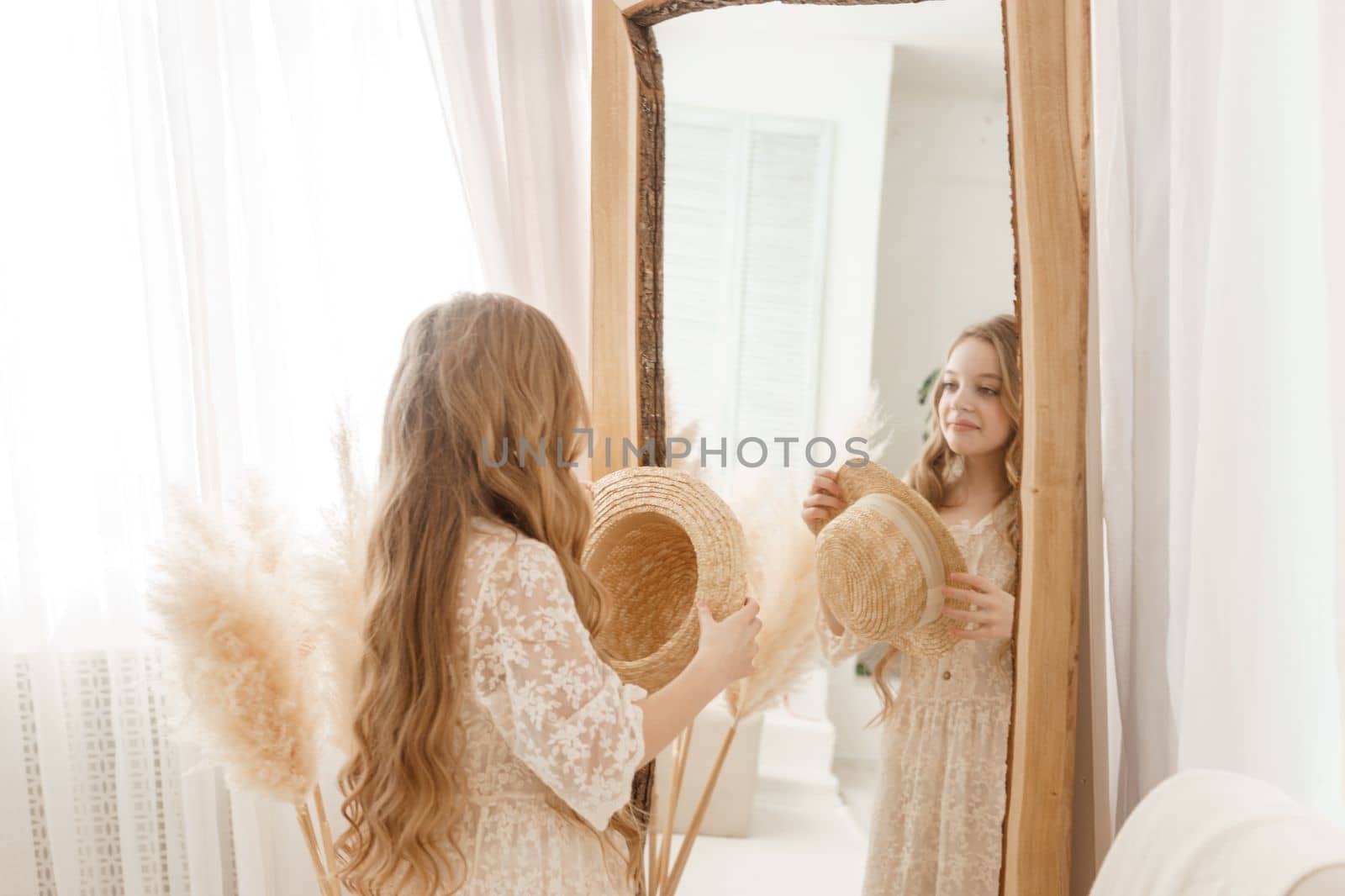 A beautiful teenage girl with long hair measures a straw hat in front of a mirror. Self-admiration of a blonde. selective focus