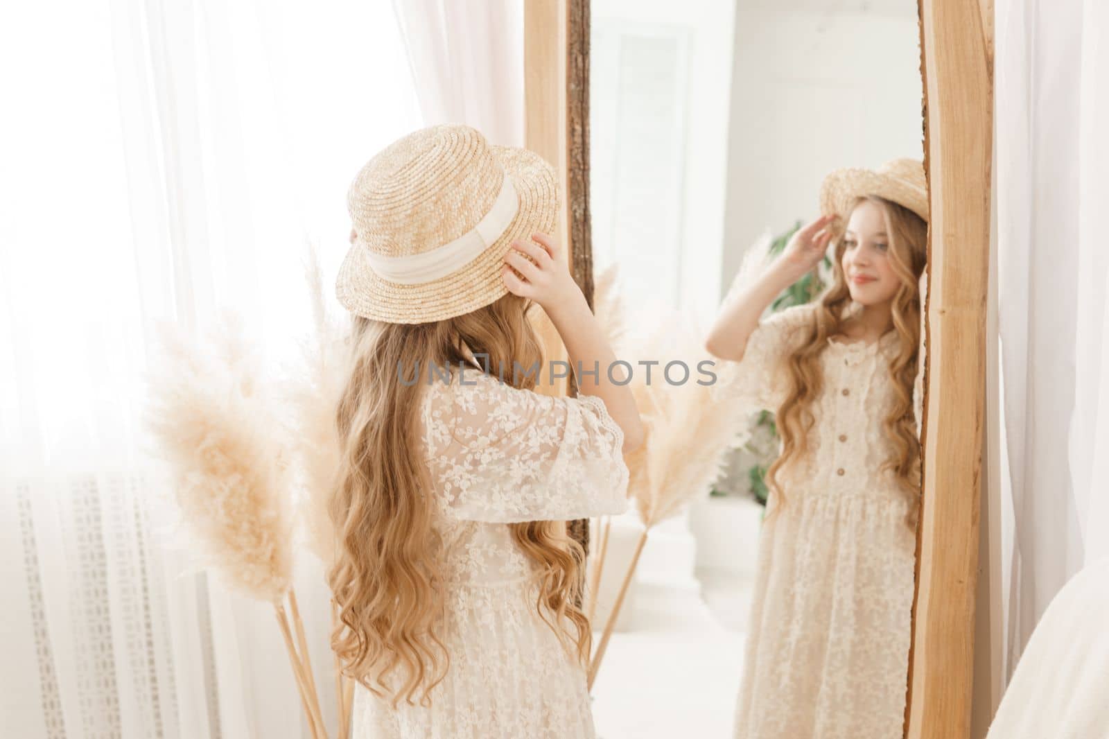 A beautiful teenage girl with long hair measures a straw hat in front of a mirror. Self-admiration of a blonde. selective focus