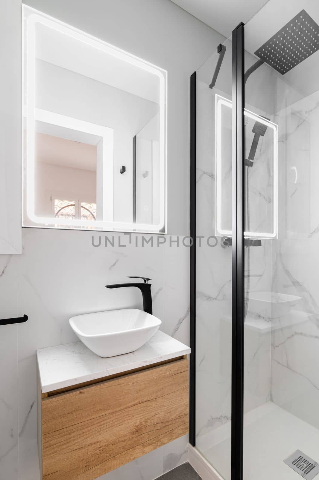 Bathroom with modern designer renovation and fittings. Vanity sink with black faucet. Walls of white granite with gray stains are beautifully illuminated by bright white light from square mirror