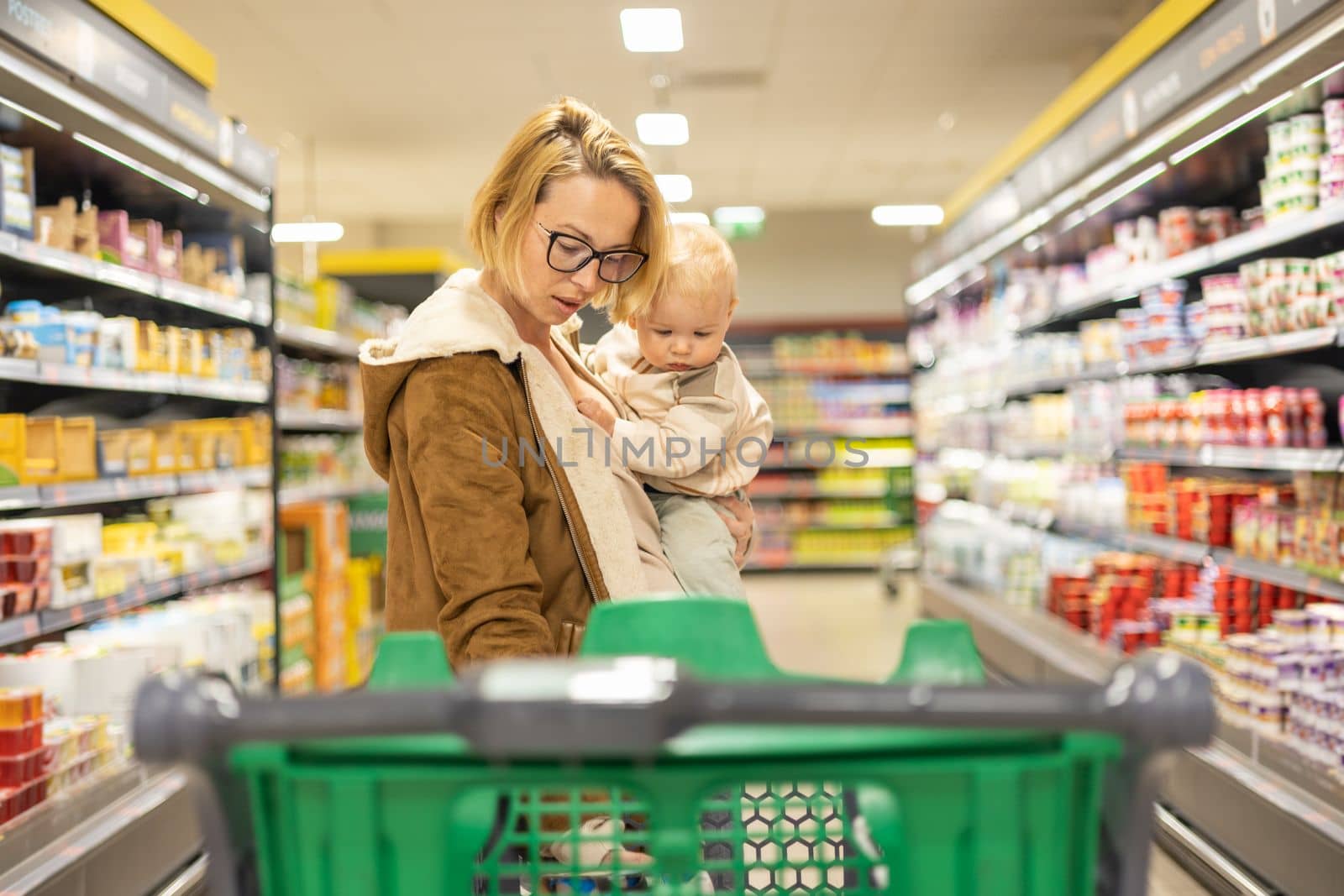 Mother shopping with her infant baby boy child, pushing shopping cart down department aisle in supermarket grocery store