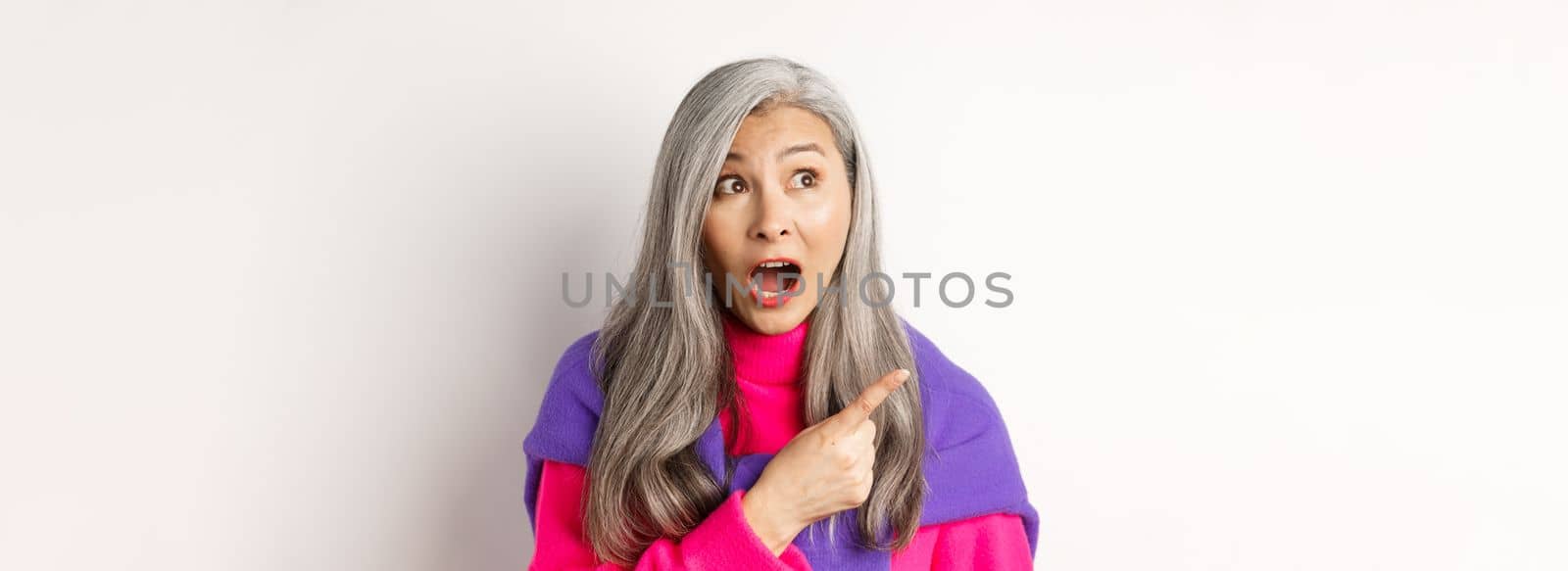 Close-up of amazed and intrigued asian senior woman pointing finger left at something cool, staring at camera promo offer, standing over white background.