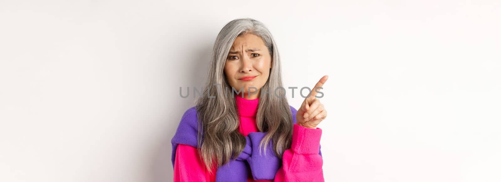 Judgy and skeptical asian senior woman grimacing displeased, pointing finger upper left corner unamused, standing over white background by Benzoix