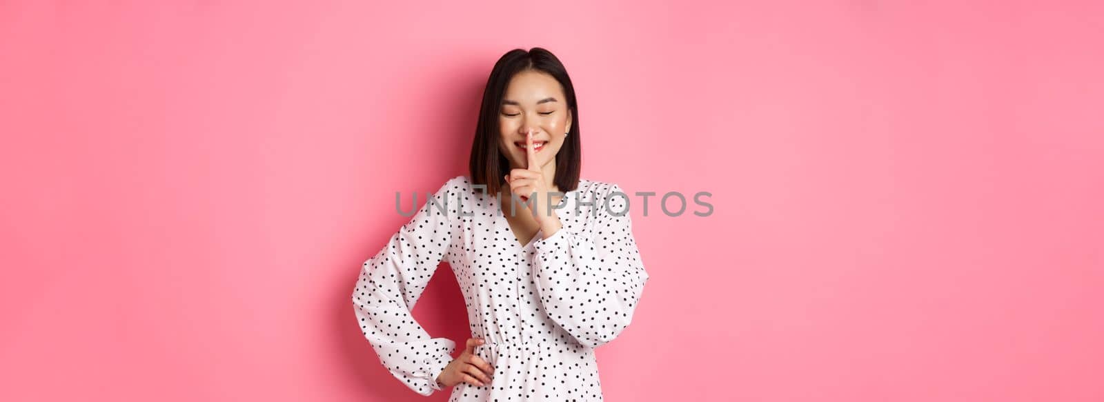 Beautiful Korean woman in trendy dress asking to keep secret, hushing with soft smile and closed eyes, standing over pink background.