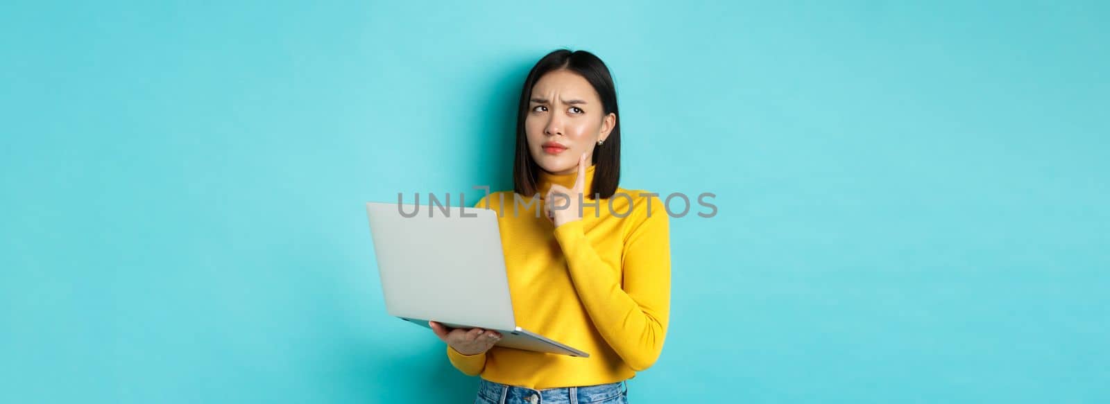Pensive asian woman working on laptop, thinking and looking away, making decision, standing against blue background.