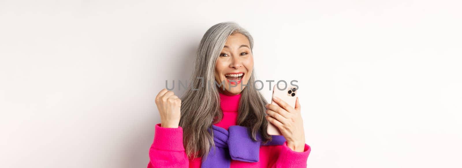 Online shopping. Close-up of cheerful asian elderly woman winning prize in internet, holding smartphone and making fist pump, standing over white background.