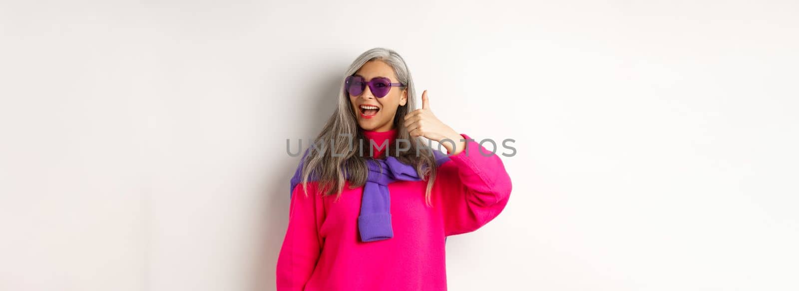 Shopping concept. Stylish asian senior woman in sunglasses and trendy outfit, showing thumb-up in approval, recommending shop, standing over white background.
