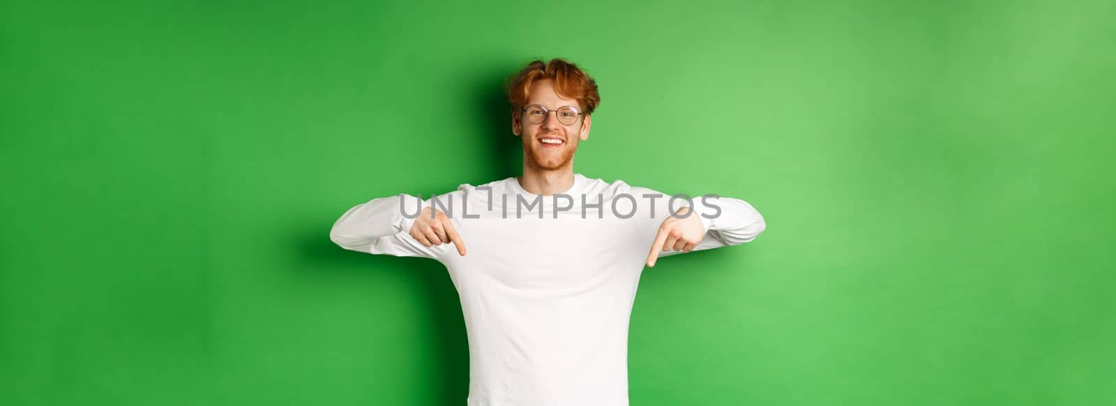 Handsome male model with red hair, wearing glasses and long-sleeve, pointing fingers down and smiling cheerful, showing promo, green background by Benzoix