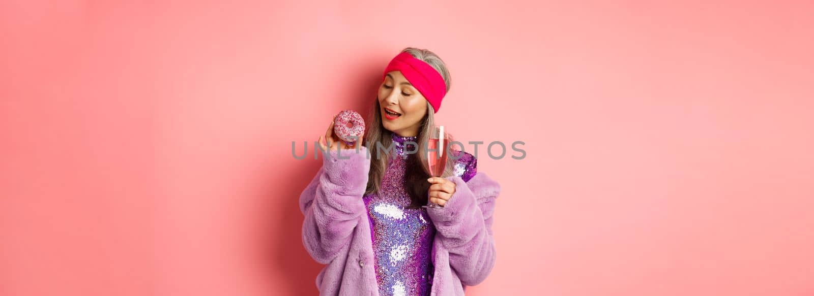 Fancy senior woman having fun, eating donut and drinking pink champagne, standing in purple faux fur coat and glittering dress, studio background.
