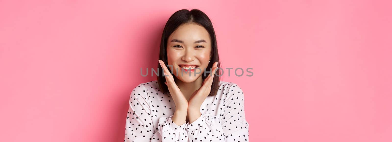 Beauty and skin care concept. Close-up of cute asian woman showing clean perfect face and smiling, looking happy at camera, standing over pink background by Benzoix