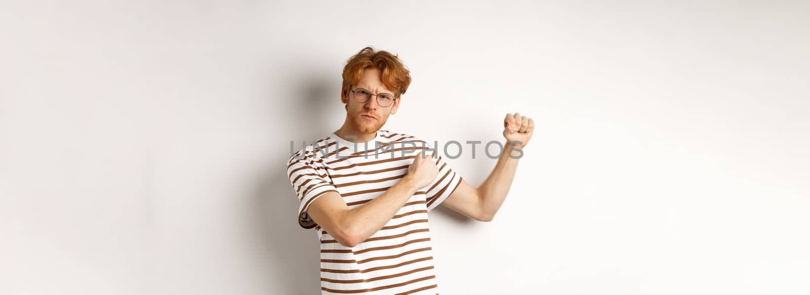 Serious and confident redhead man in glasses raising fists, ready for fight, staring angry at camera while shadow boxing, standing over white background.