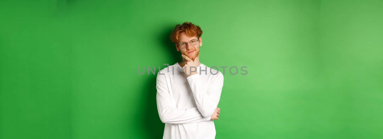 Intrigued young man with red hair, wearing glasses, looking at camera pleased and thoughtful, having an idea, standing over green background.