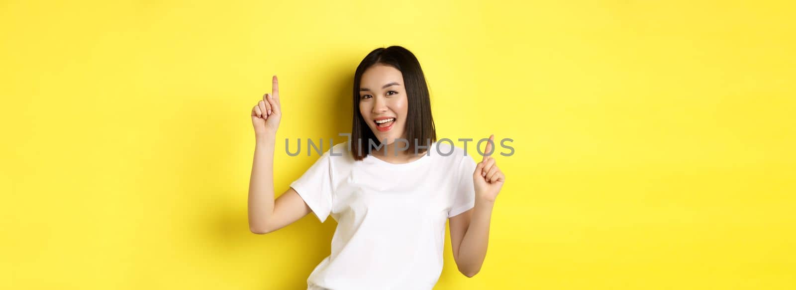 Happy asian woman dancing and having fun, posing in white t-shirt against yellow background.