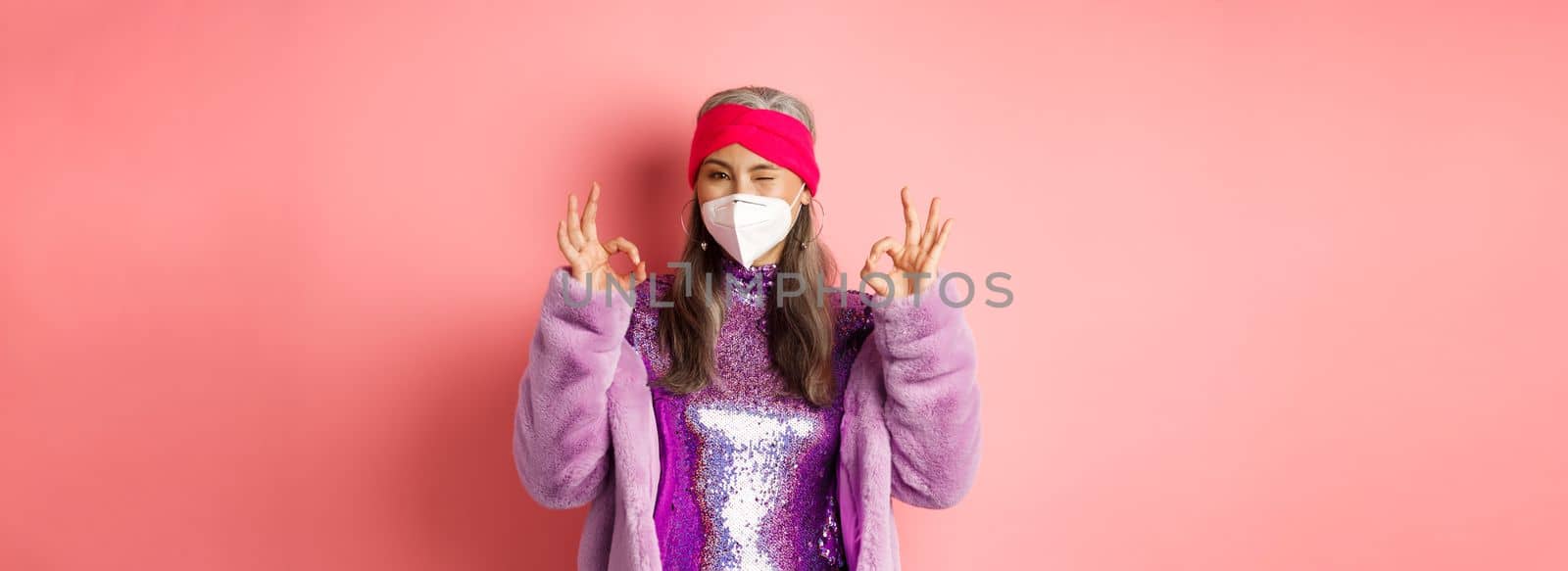 Covid-19, pandemic and fashion concept. Awesome asian grandmother wearing stylish disco dress and respirator, showing okay signs and winking, no proble gesture.