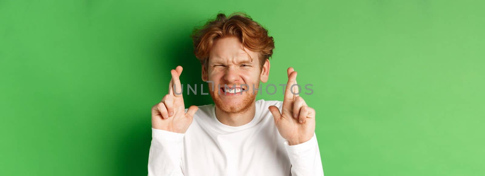 Hopeful caucasian man looking worried, waiting for important results and making wish with fingers crossed, standing over green background.