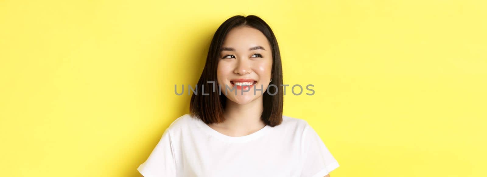 Close up of good-looking asian woman in white t-shirt, smiling and glancing left with pleased face, standing over yellow background by Benzoix