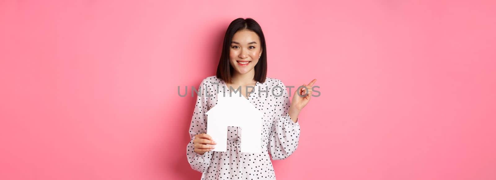Real estate. Excited asian woman showing paper house model, pointing left at copy space, standing over pink background by Benzoix