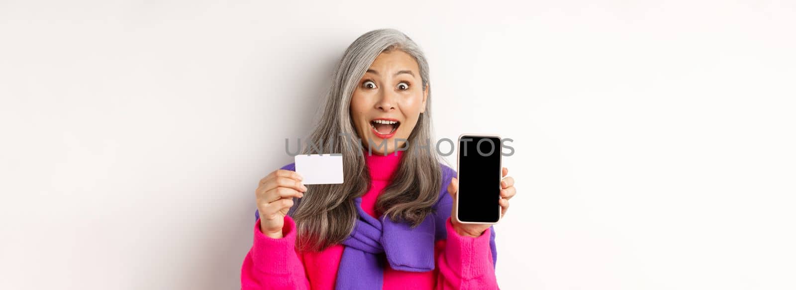 Online shopping. Closeup of amazed asian senior woman showing blank mobile screen and plastic credit card, looking happy, standing over white background.