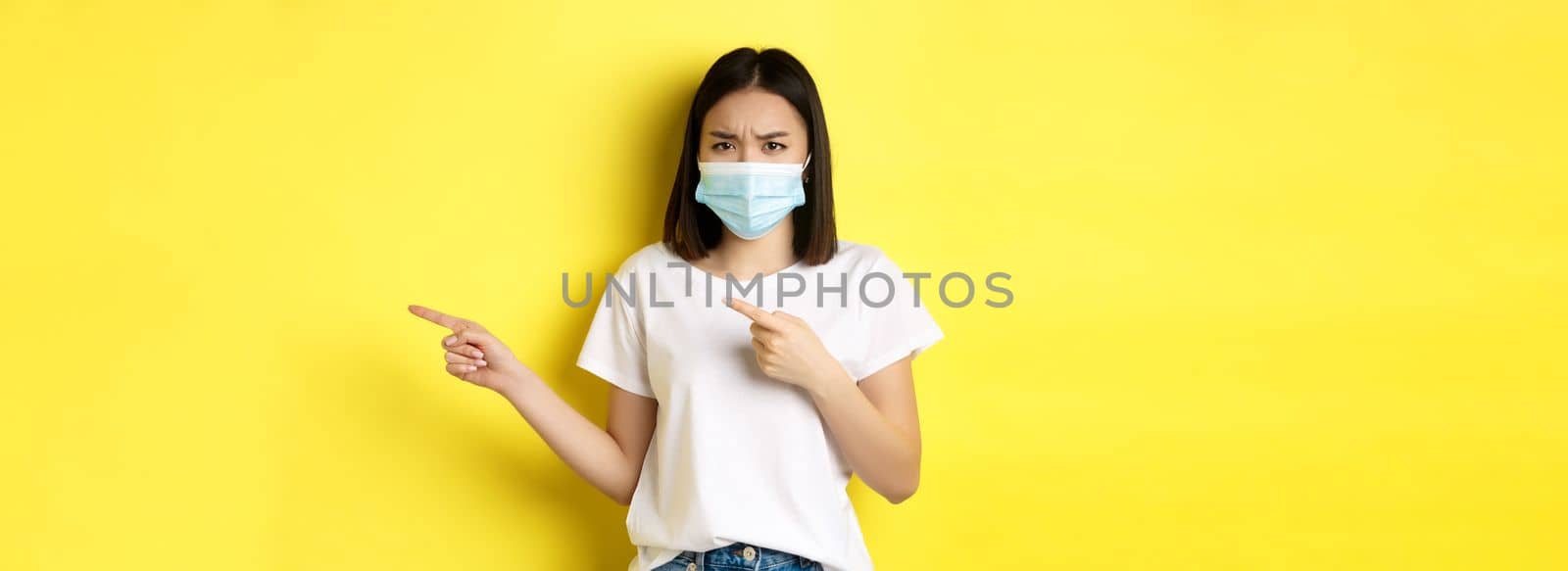 Covid-19, pandemic and social distancing concept. Disappointed asian girl in medical mask, frowning upset and pointing fingers right at logo, standing over yellow background by Benzoix