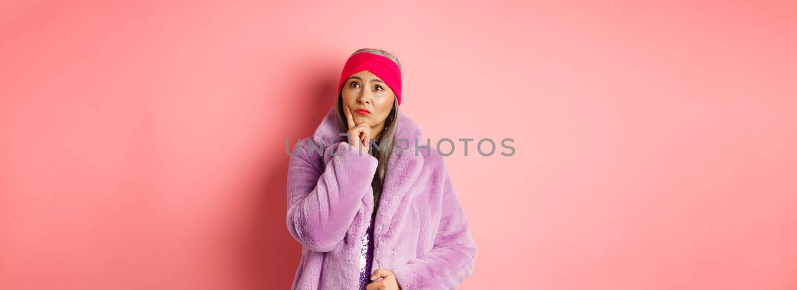 Funky old asian woman looking thoughtful and perplexed, standing in fake fur coat and thinking, making decision, standing on pink background by Benzoix