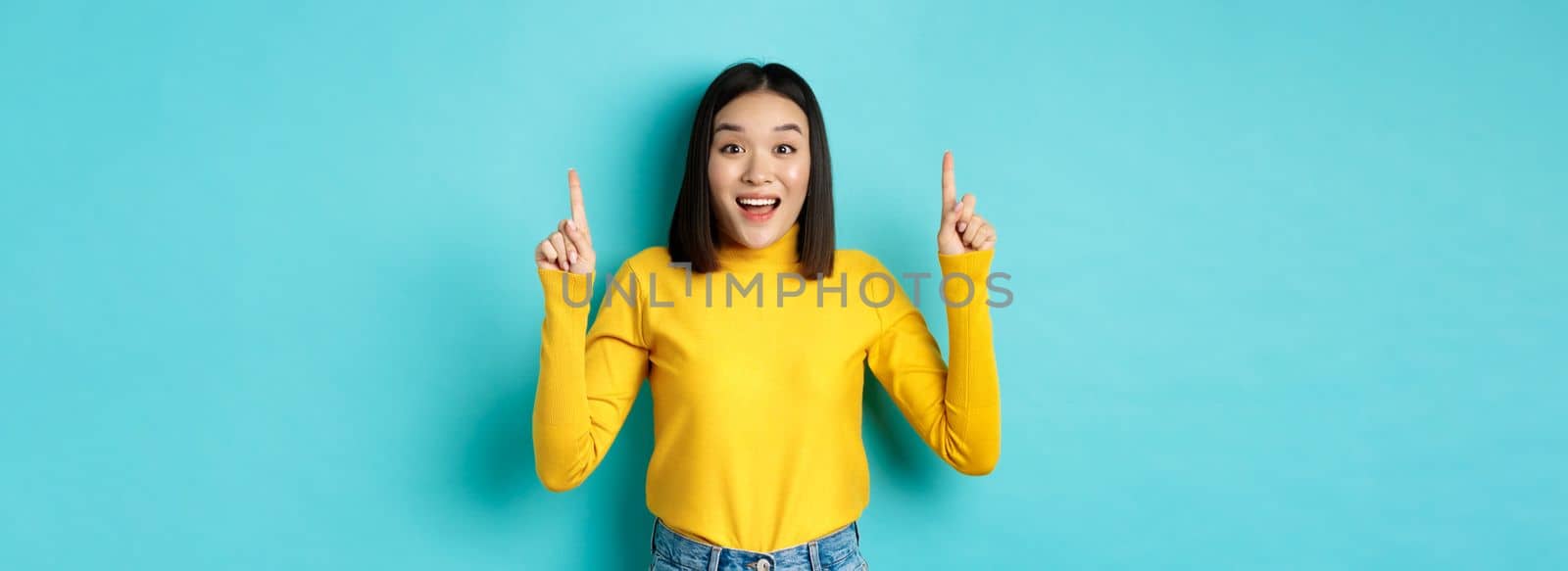 Shopping concept. Impressed young korean woman showing advertisement on top, pointing fingers up and gasping amazed, saying wow, standing over blue background.