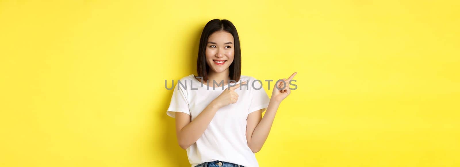 Beauty and fashion concept. Beautiful asian woman in white t-shirt pointing fingers left, standing over yellow background by Benzoix