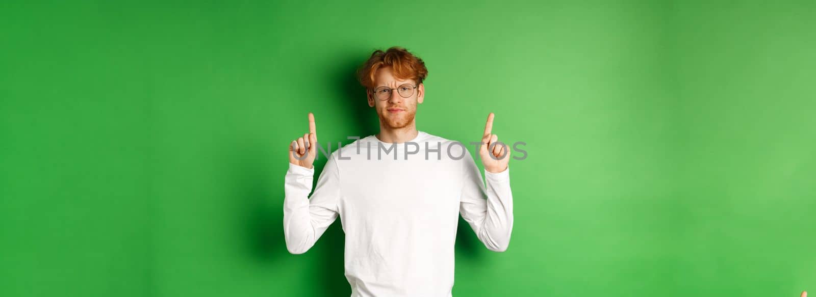 Displeased young man with red hair and glasses, pointing fingers up and frowning doubtful, feeling disappointed, standing over green background.