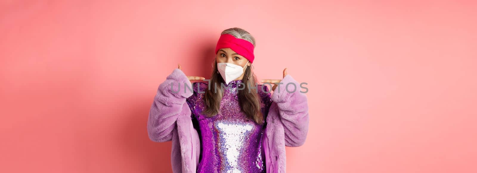 Covid-19, pandemic and fashion concept. Fashionable asian grandmother wearing glittering party dress and respirator, pointing at mask, pink background.