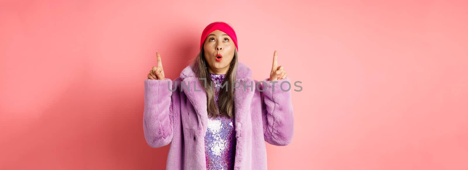 Impressed asian elderly woman in stylish fake-fur coat checking out promo, looking and pointing fingers up, standing over pink background.