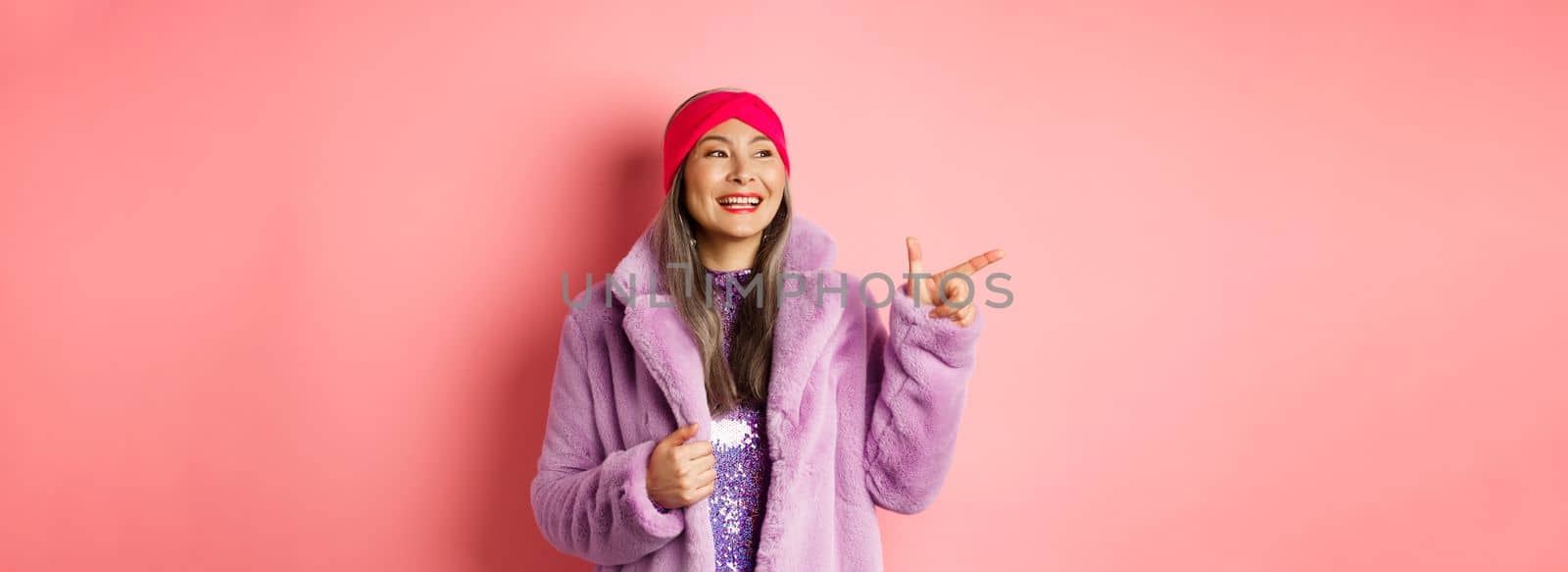 Happy senior asian woman smiling, pointing finger left and looking satisfied, checking out special promotion, pink background.
