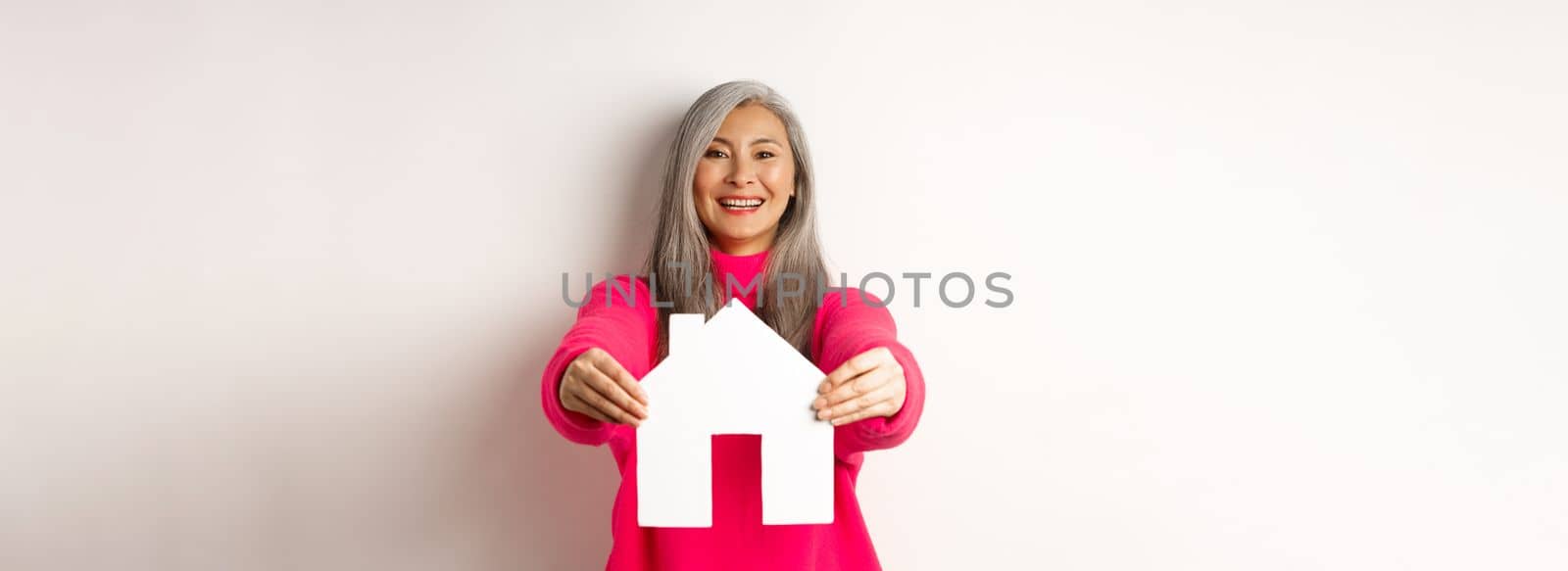 Real estate. Happy korean middle-aged woman with grey hair, showing paper house model and smiling, standing in pink sweater over white background.