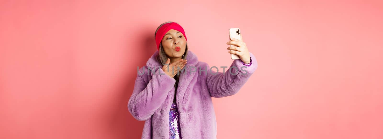 Fashion concept. Beautiful and trendy asian mature woman in faux fur coat with party dress, taking selfie, sending kiss to smartphone camera, posing against pink background.