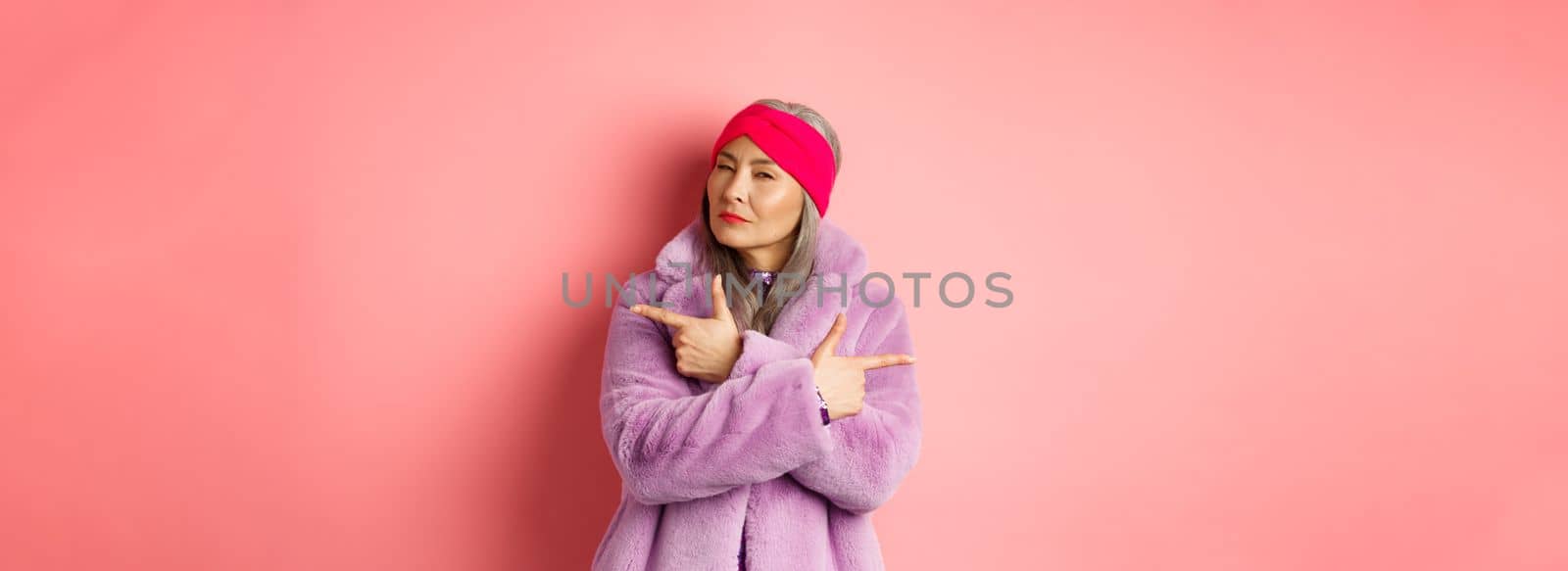 Fashion and shopping concept. Stylish asian mature woman making decision, squinting thoughtful while pointing fingers sideways, showing two variants on pink background.