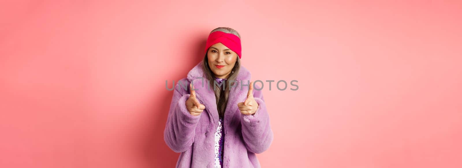 Fashion and shopping concept. Cool asian senior woman in stylish fake fur coat pointing fingers at camera, asking you to check out promo offer, standing over pink background.