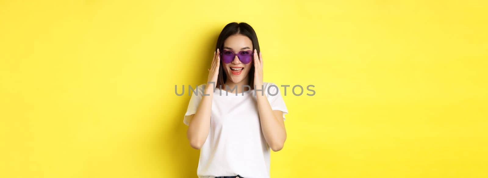 Fashion and lifestyle concept. Stylish asian woman trying new sunglasses in heart-shape, going on vacation, smiling happy at camera, standing over yellow background.