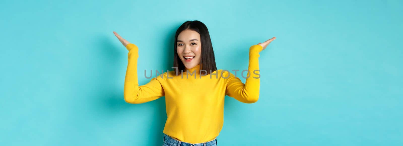 Beauty and fashion concept. Stylish beautiful asian girl demonstrate promotion, raising hands up and smiling, showing something on blue background.