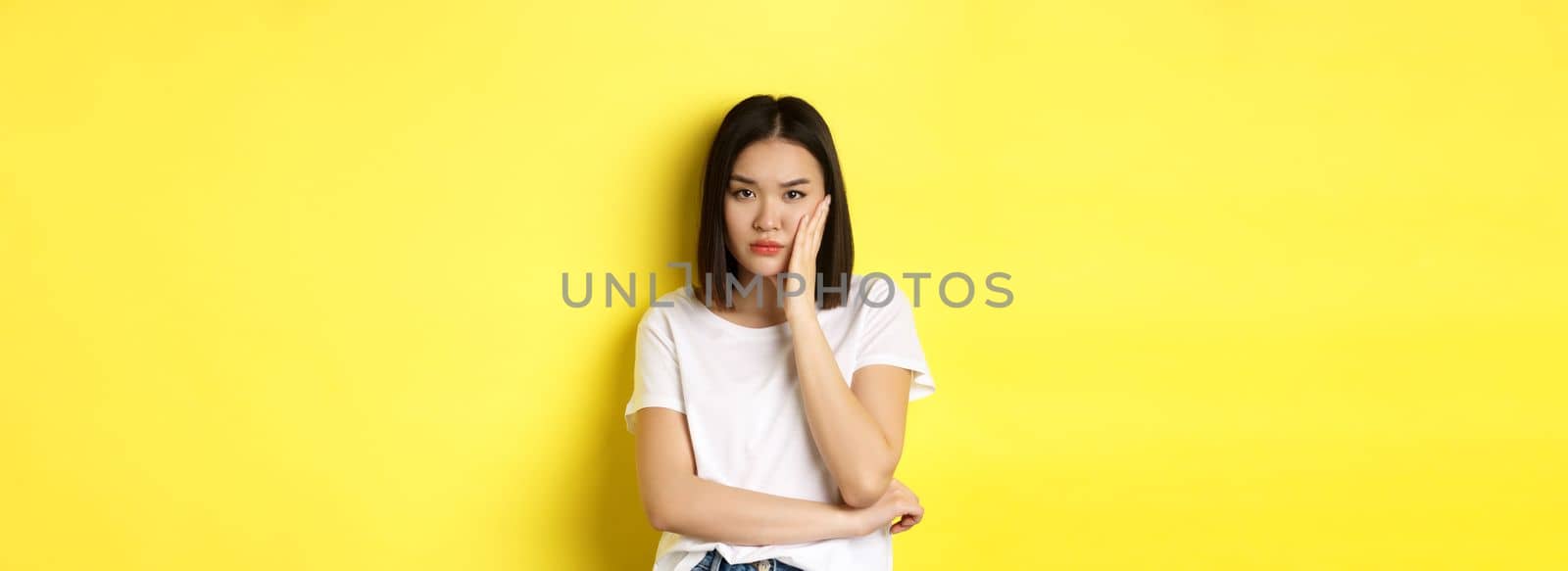 Bored and annoyed asian woman tired of listening, looking skeptical at camera, standing over yellow background.